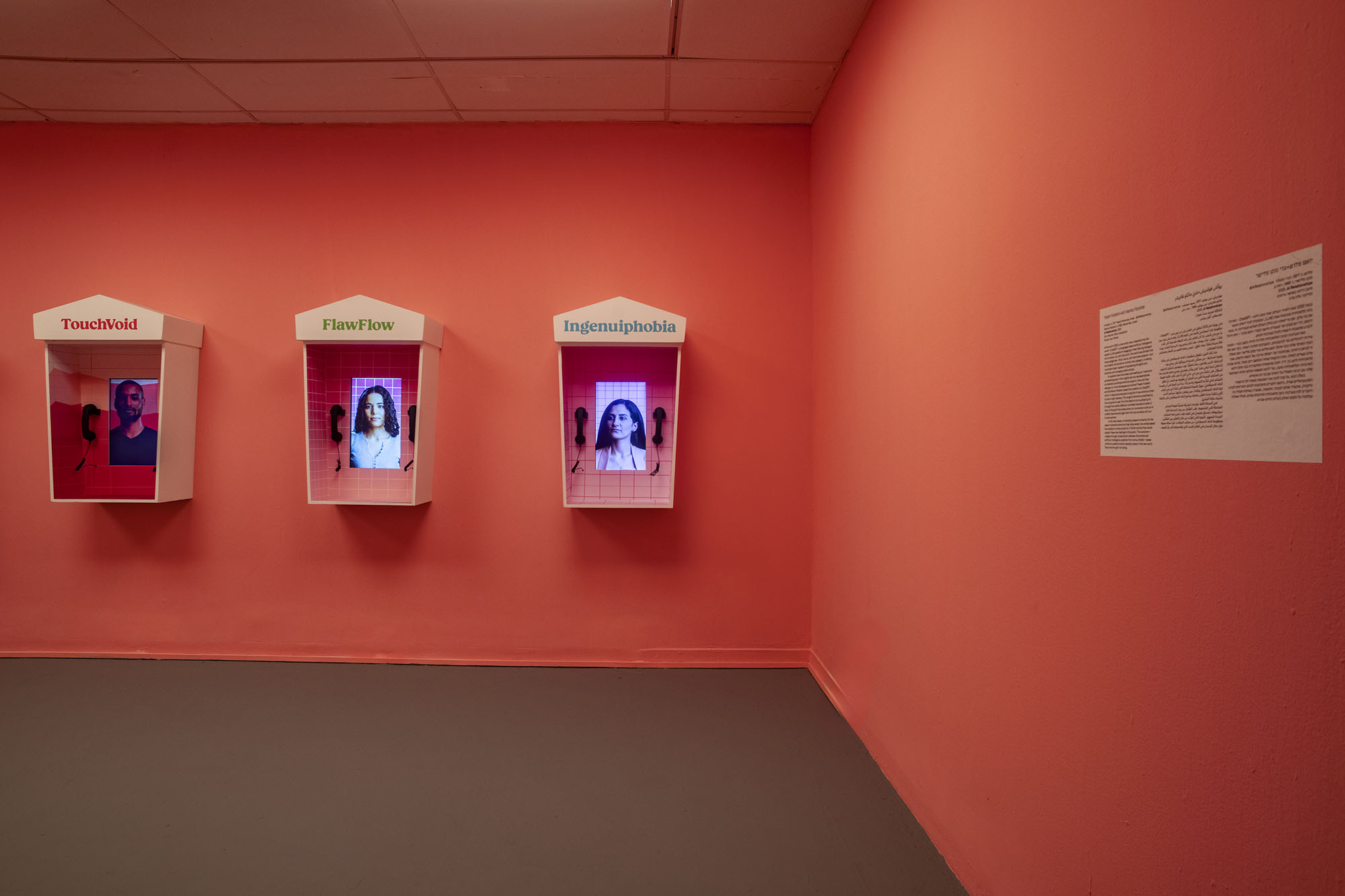 AI Relationships — gallery installation: white payphone kiosks named TouchVoid, FlawFlow and Ingenuiphobia on a coral wall, each screen holding a person, with the exhibition wall text nearby