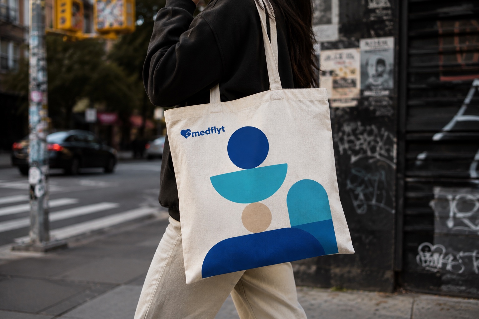 Medflyt tote bag on a city street — the brand's caregiver-and-patient shapes in blues and sand on natural canvas